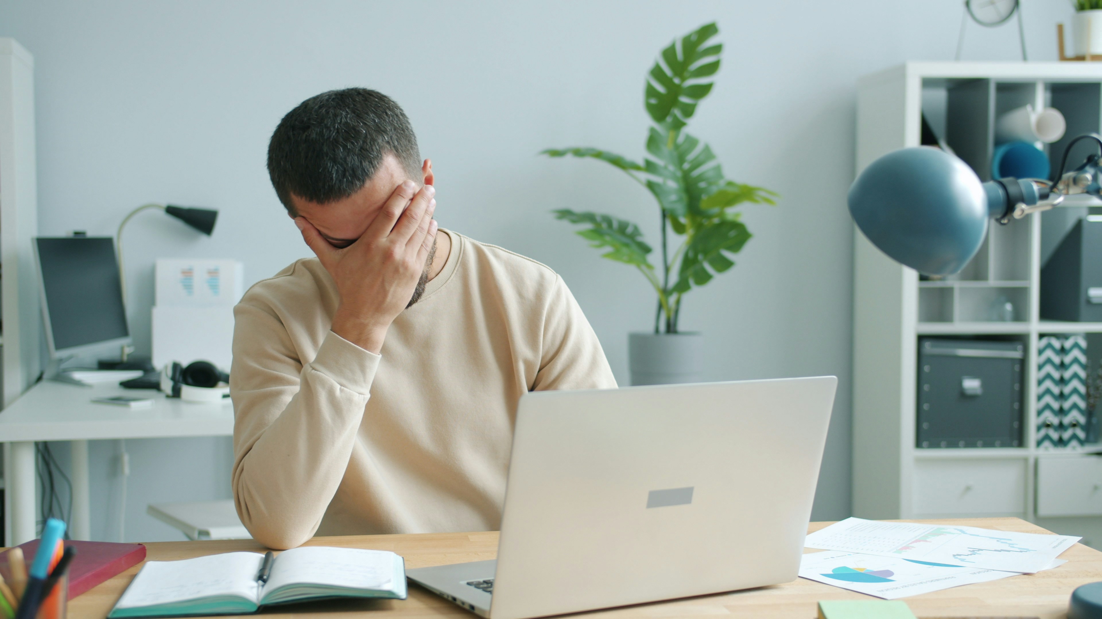 A person sitting in front of an open laptop and notebooks, holding their palm against their face in frustration.
